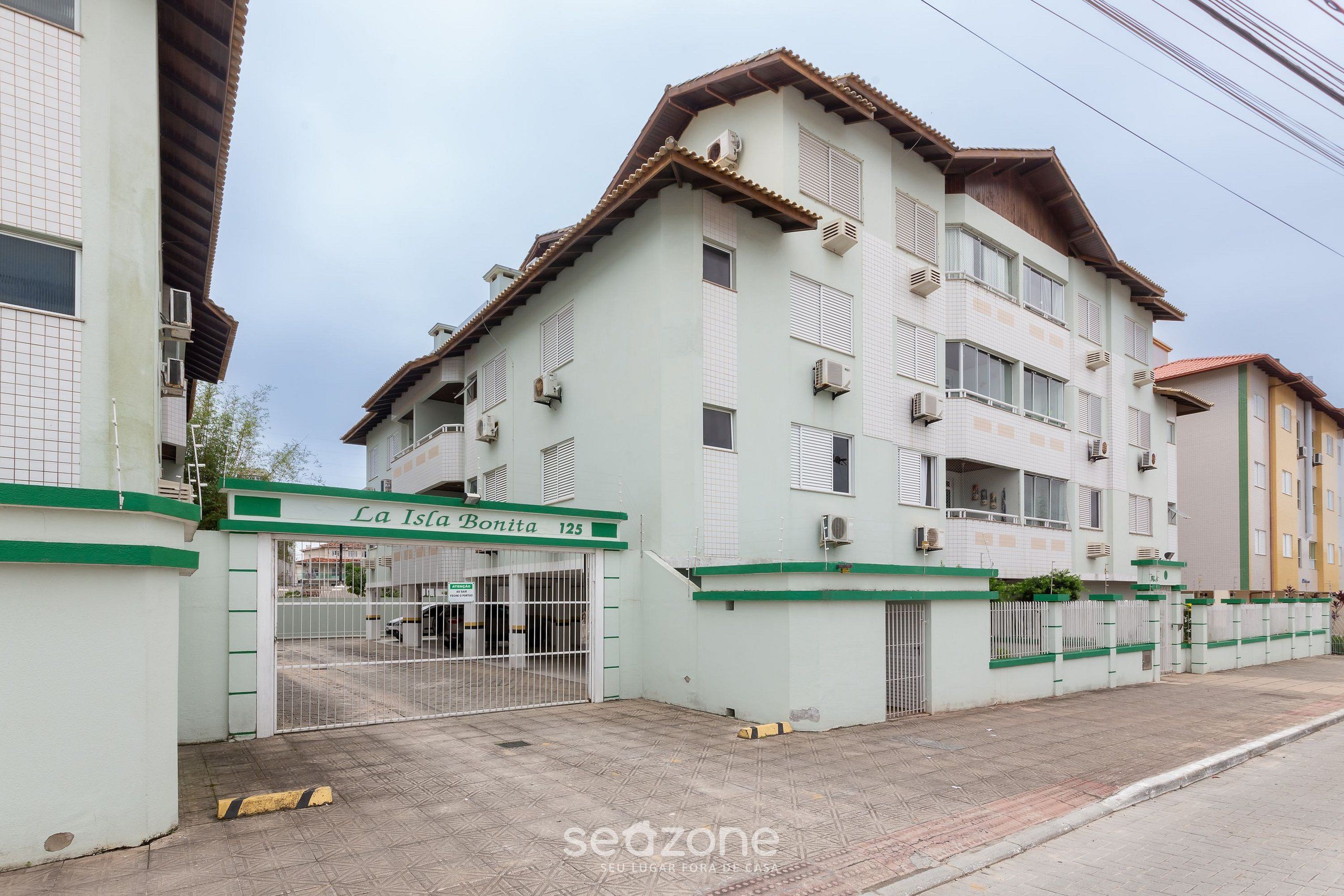 Un edificio residencial de varios pisos con entrada protegida por un portón, unidades de aire acondicionado y un cartel con el nombre del edificio y el número de la unidad.