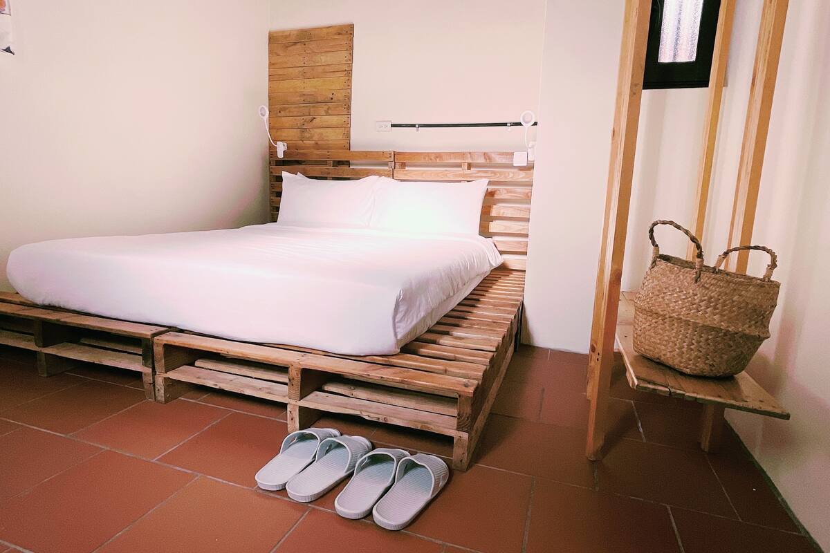 A single bed with a wooden headboard, a white mattress, and a pair of slippers on the floor.