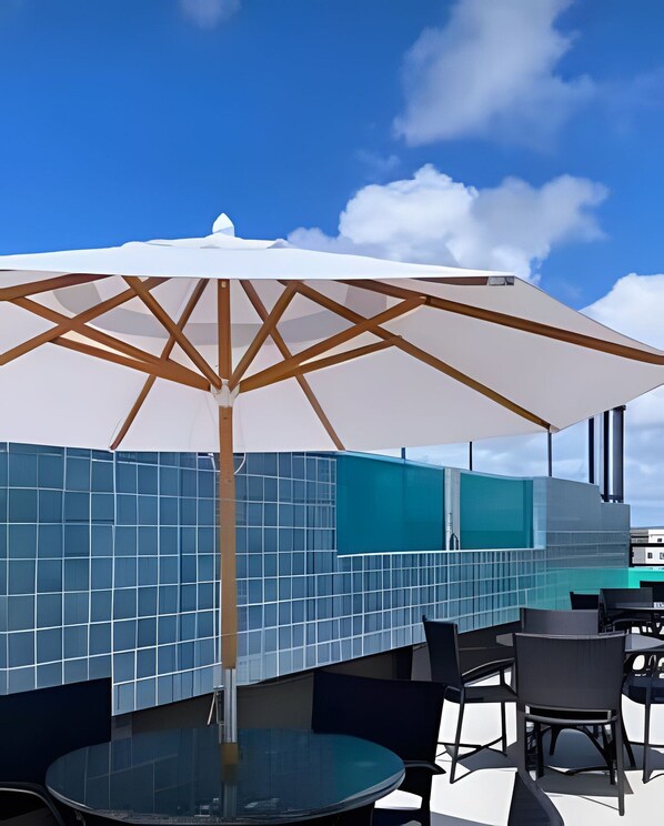 A rooftop bar with a glass table, chairs, and a white umbrella. - Varandas Porto de Galinhas (Ipojuca)