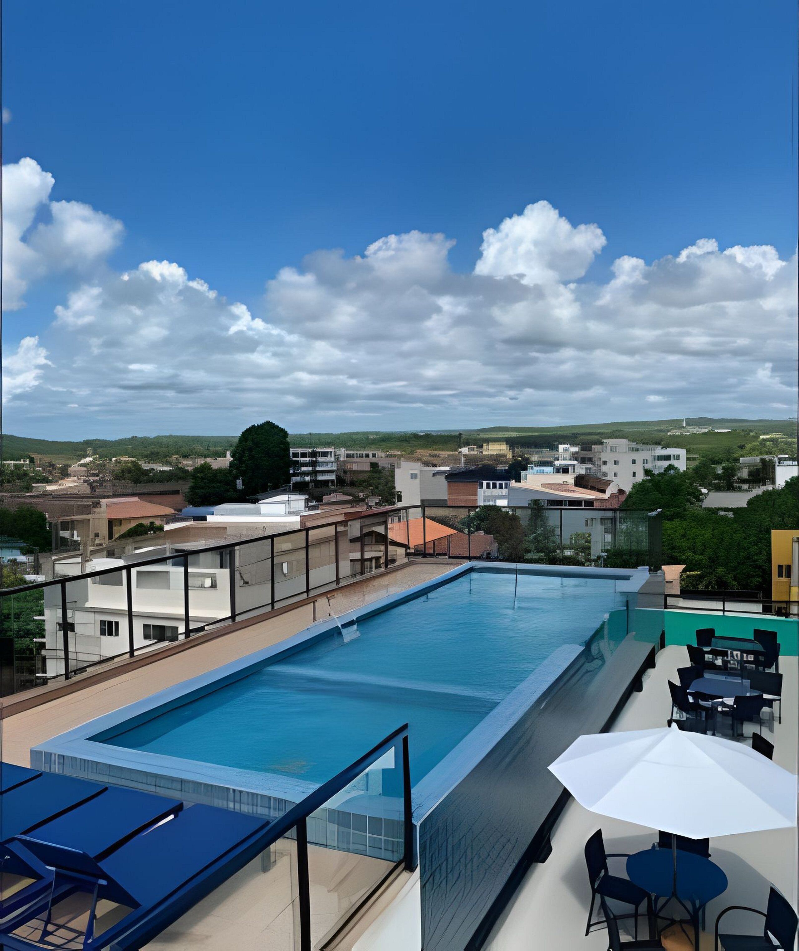 Piscina en la azotea con barandilla de cristal, mobiliario exterior y vistas de edificios y colinas circundantes.