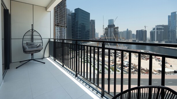 A balcony with a view of a cityscape, a hanging chair, and a railing. - KeyRock Living - Elegant Lake View Apt (Dubai)