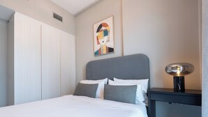 A neatly made bed with white linens and two gray pillows, a gray headboard, a wall-mounted abstract painting, and a bedside table with a lamp. - KeyRock Living - Elegant Lake View Apt (Dubai)