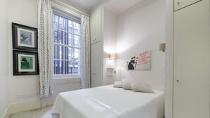 Een slaapkamer met een bed, een raam met gordijnen, een wandlamp en ingelijste foto's.