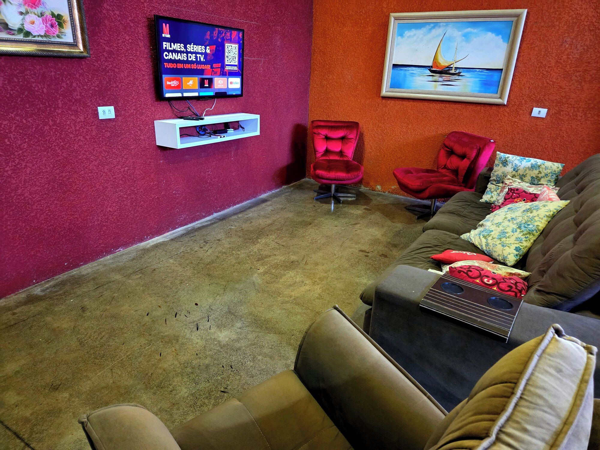 Un salon avec un mur rouge, un téléviseur fixé au-dessus d’un meuble de télévision blanc, un fauteuil rouge, un canapé marron avec des coussins à motifs et un tableau encadré représentant un voilier.