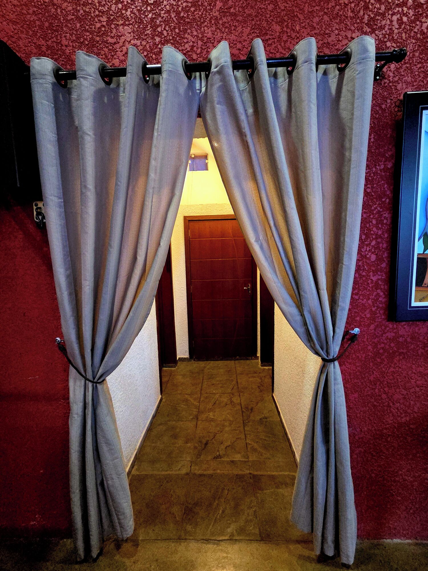 Um corredor com parede vermelha texturizada, uma porta de madeira no fim e cortinas cinzas amarradas no alto.