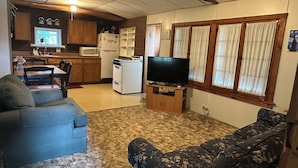 Una sala de estar acogedora con televisor, dos sofás, una cocina con refrigerador, microondas y estufa, y un comedor con sillas y mesa.
