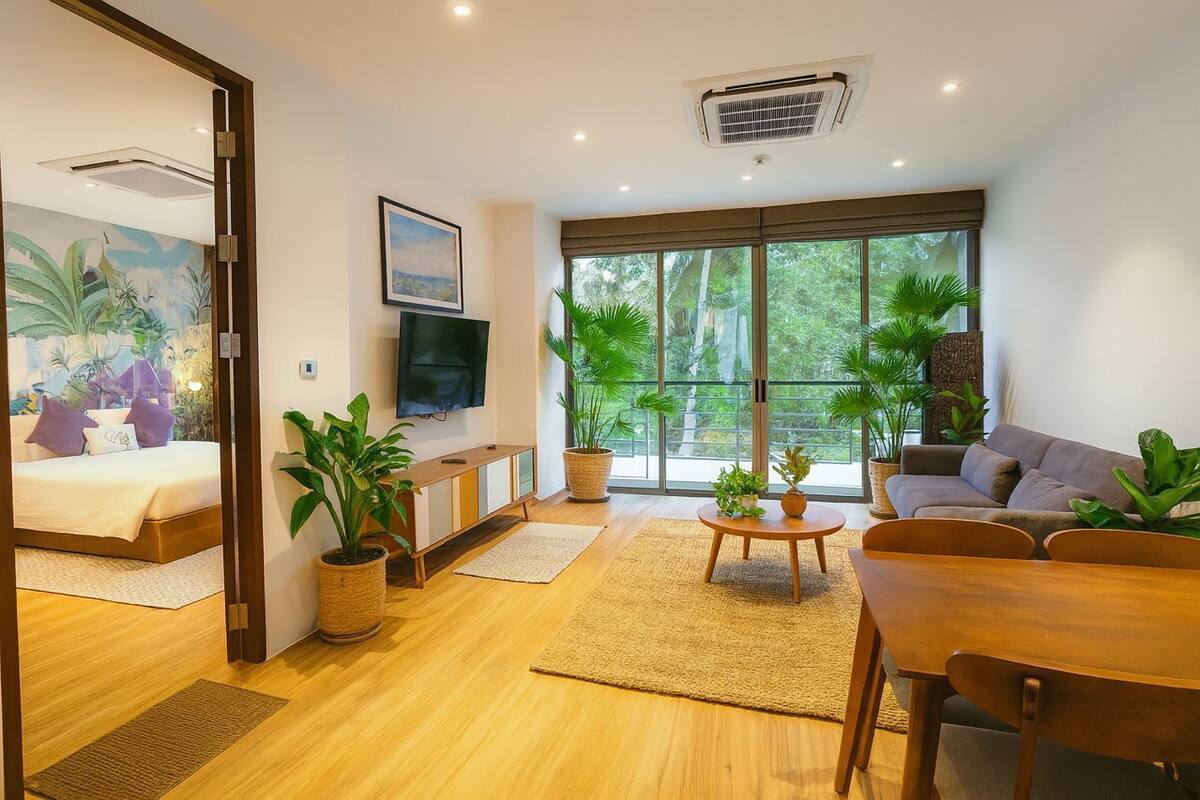 Un soggiorno moderno con un divano, un tavolo da pranzo e una TV. Sono presenti piante in vaso e un'ampia finestra con vista sugli alberi.
