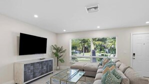A modern living room with a flat-screen TV, a glass coffee table, a beige sofa, and a large window with blinds. - Walking distance to the beach & close to downtown! (Boca Raton)