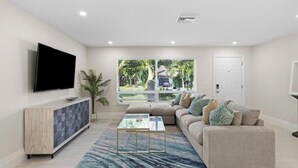 A modern living room with a flat-screen TV, a sofa, a coffee table, and a view of the outdoors. - Walking distance to the beach & close to downtown! (Boca Raton)