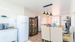 A modern kitchen with white cabinets, a white refrigerator, and a microwave. There is a dining area with a glass table and beige chairs. - Beautiful apt 750 m From the Beach of Ingleses Lgr204 (Florianópolis)