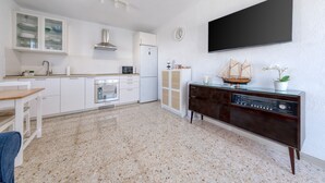 Een moderne keuken met witte kastjes, een ingebouwde oven en een magnetron. Er is een eettafel met stoelen, een televisie aan de muur en een decoratief model schip op een kast.