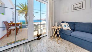Een woonkamer met een blauwe bank, een houten tafel en uitzicht op zee door grote glazen deuren.