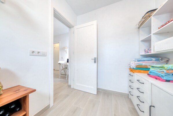 Un couloir avec une porte blanche, une étagère en bois avec des bouteilles et un meuble de rangement contenant des vêtements pliés.