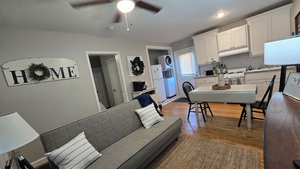 A modern kitchen with white cabinets, a dining area with a table and chairs, and a living room with a sofa and a ceiling fan.