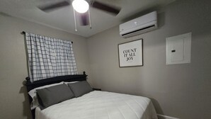 A bedroom with a bed, a ceiling fan, an air conditioning unit, a framed quote on the wall, and a door.