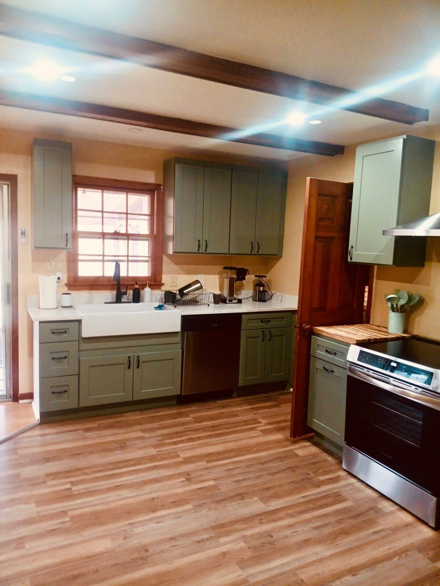 A kitchen with wooden flooring, green cabinets, a stainless steel stove, and a window with blinds.