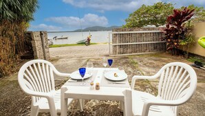 Mesa posta para dois com cadeiras de plástico branco, vista para um lago com barco e uma moto, e um muro de pedra com vegetação.