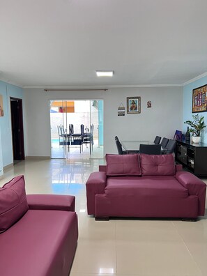 A spacious living area with a large sofa and a dining table, a sliding glass door leading to an outdoor patio, and a small decorative shelf with a plant.