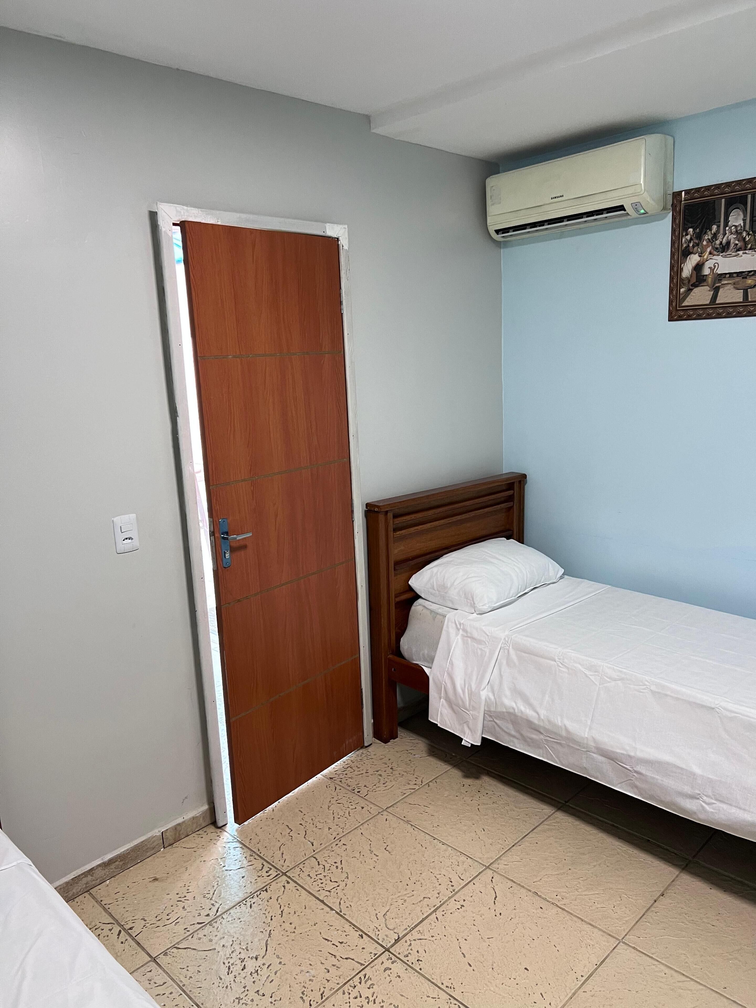 A bedroom with a wooden door, a bed with white bedding, a wall-mounted air conditioner, and a tiled floor.