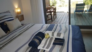 Un lit bien fait, avec une literie à rayures et des coussins décoratifs, une table de chevet avec une lampe, et une vue sur une terrasse extérieure avec des chaises et une table.
