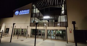 Un hotel llamado 'Adonis' con un diseño arquitectónico moderno, que destaca un cartel llamativo y una entrada de cristal.