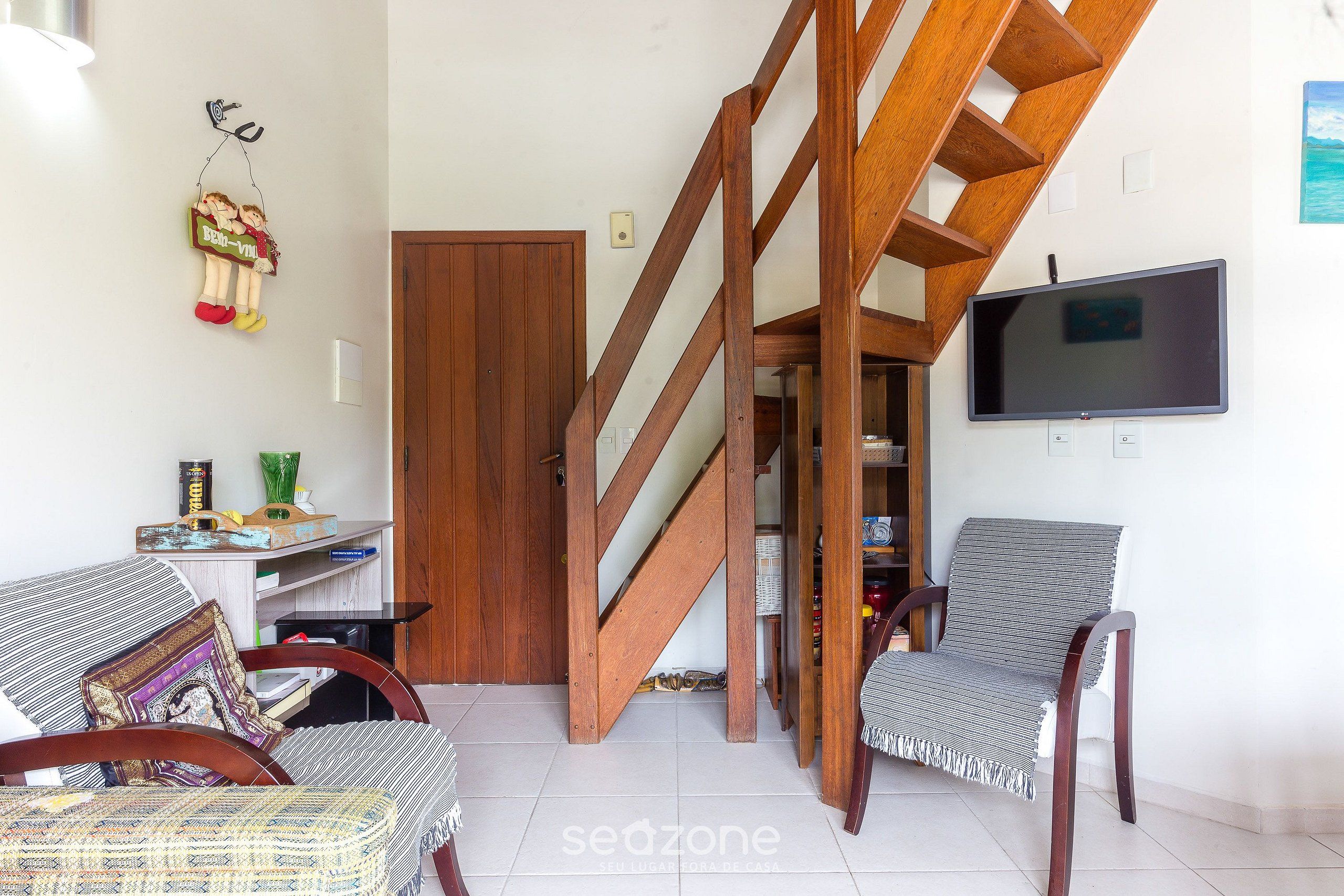 Una sala de estar con una escalera de madera, un televisor montado en la pared y dos sillones.