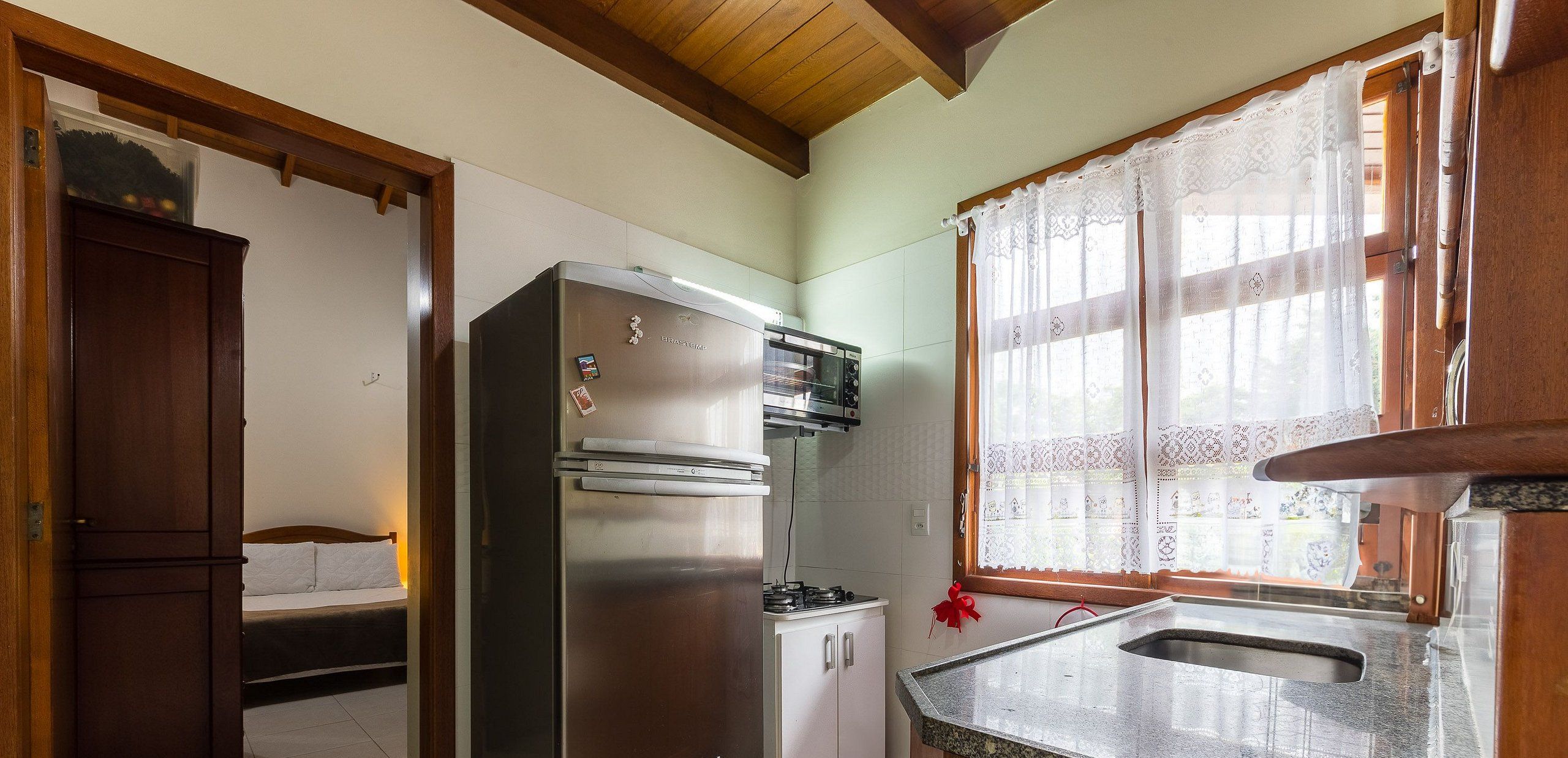 A compact kitchen with a refrigerator, oven, and microwave. There's a bed in the adjacent room and a window with lace curtains.