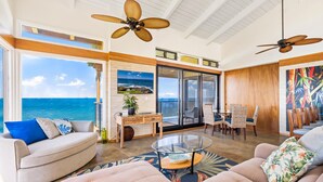 A spacious living room with a large window overlooking the ocean, a ceiling fan, and a dining area with wicker chairs.