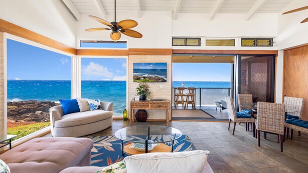 A spacious living area with a large window overlooking the ocean, a ceiling fan, and a dining area with wicker furniture.