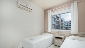Una habitación limpia y moderna con dos camas individuales, una mesita de noche pequeña, una ventana con cortinas y un equipo de aire acondicionado en la pared.