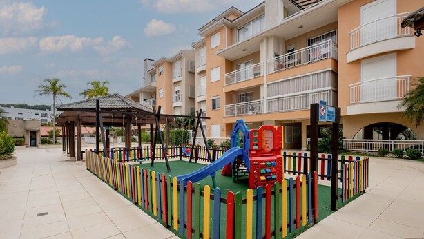 Zona de juegos con tobogán y estructuras para escalar, rodeada por una valla de colores, frente a un edificio de varios pisos con balcones.