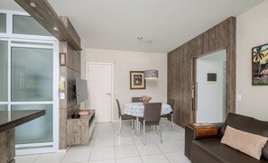 A modern living room with a brown sofa, a dining table with chairs, and a kitchen area with a fridge and a TV.