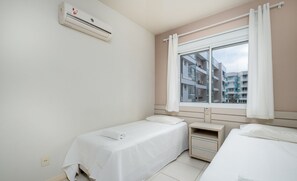 A clean, modern room with two single beds, a small bedside table, a window with curtains, and an air conditioning unit on the wall.