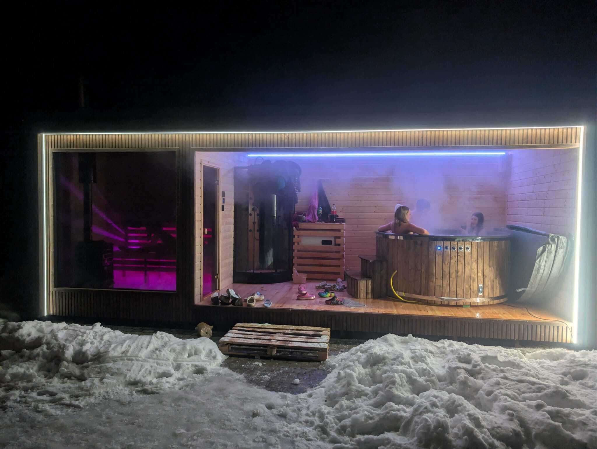 Una sauna con dos personas adentro, un banco de madera afuera y un montículo de nieve adelante.