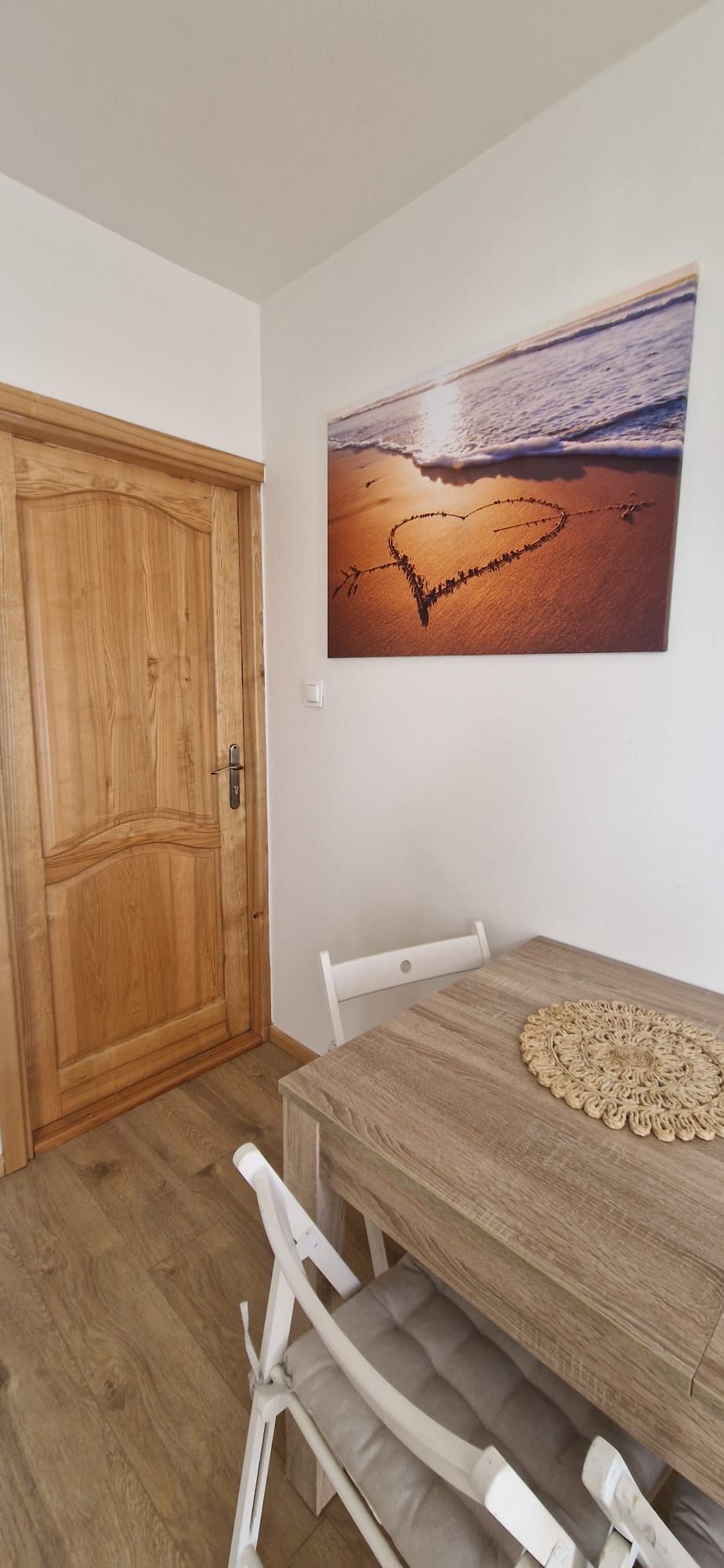 Un comedor con una mesa de madera, sillas blancas y un cuadro en la pared que muestra una playa.