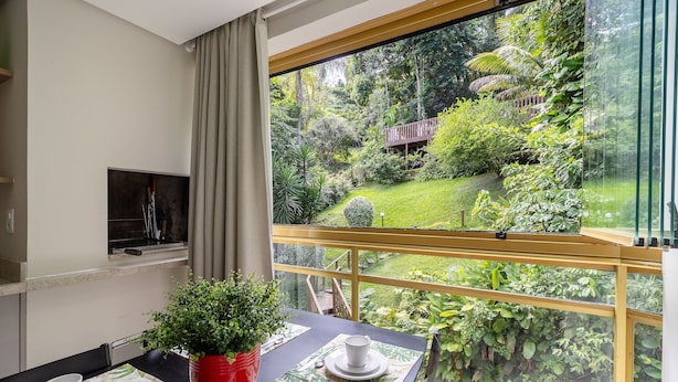 A dining area with a table set for two, a potted plant, and a view of a lush garden through a large window. - Sea View Apt 240m From the Beach in Jurere Ido207 (Florianópolis)