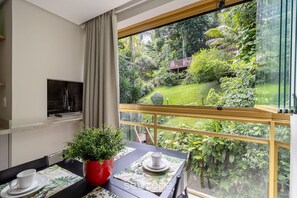 Ein Zimmer mit Esstisch, gedeckt für zwei Personen, einer roten Topfpflanze und Blick auf einen üppigen Garten durch ein großes Fenster.