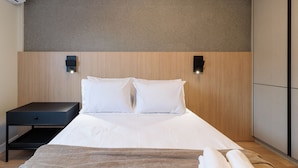 Una cama bien tendida con sábanas blancas, una mesita de noche oscura con cajón y dos luces fijadas a la pared.