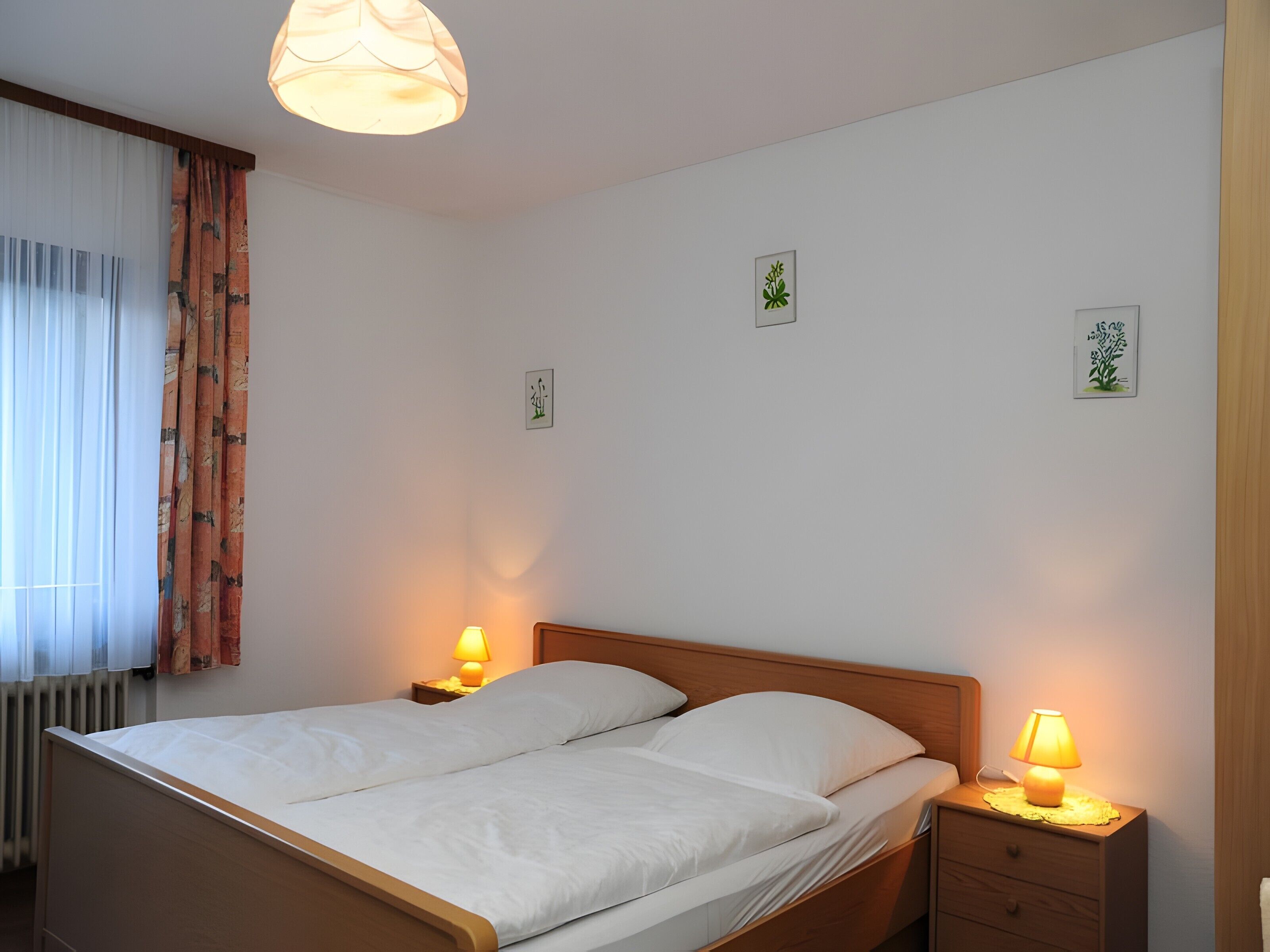 Een tweepersoonsbed met twee nachtkastjes, elk voorzien van een lamp, een raam met gordijnen en wanddecoratie.