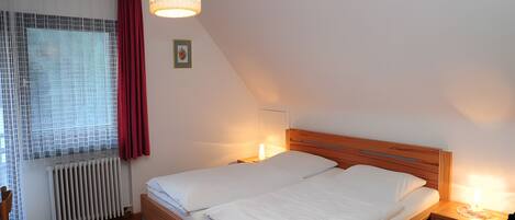 Quarto com cama de madeira, mesa de cabeceira, luminária pendente, radiador e janela com cortinas.