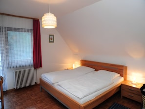 A bedroom with a wooden bed, a bedside table, a nightstand, a hanging light fixture, a radiator, and a window with curtains. - Apartment 'Nr 2' with Shared Terrace, Shared Garden and Wi-Fi (Simonswald)