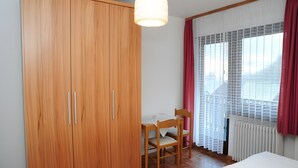 A bedroom with a wooden wardrobe, a wooden chair, a bed, a hanging light fixture, and a window with curtains.