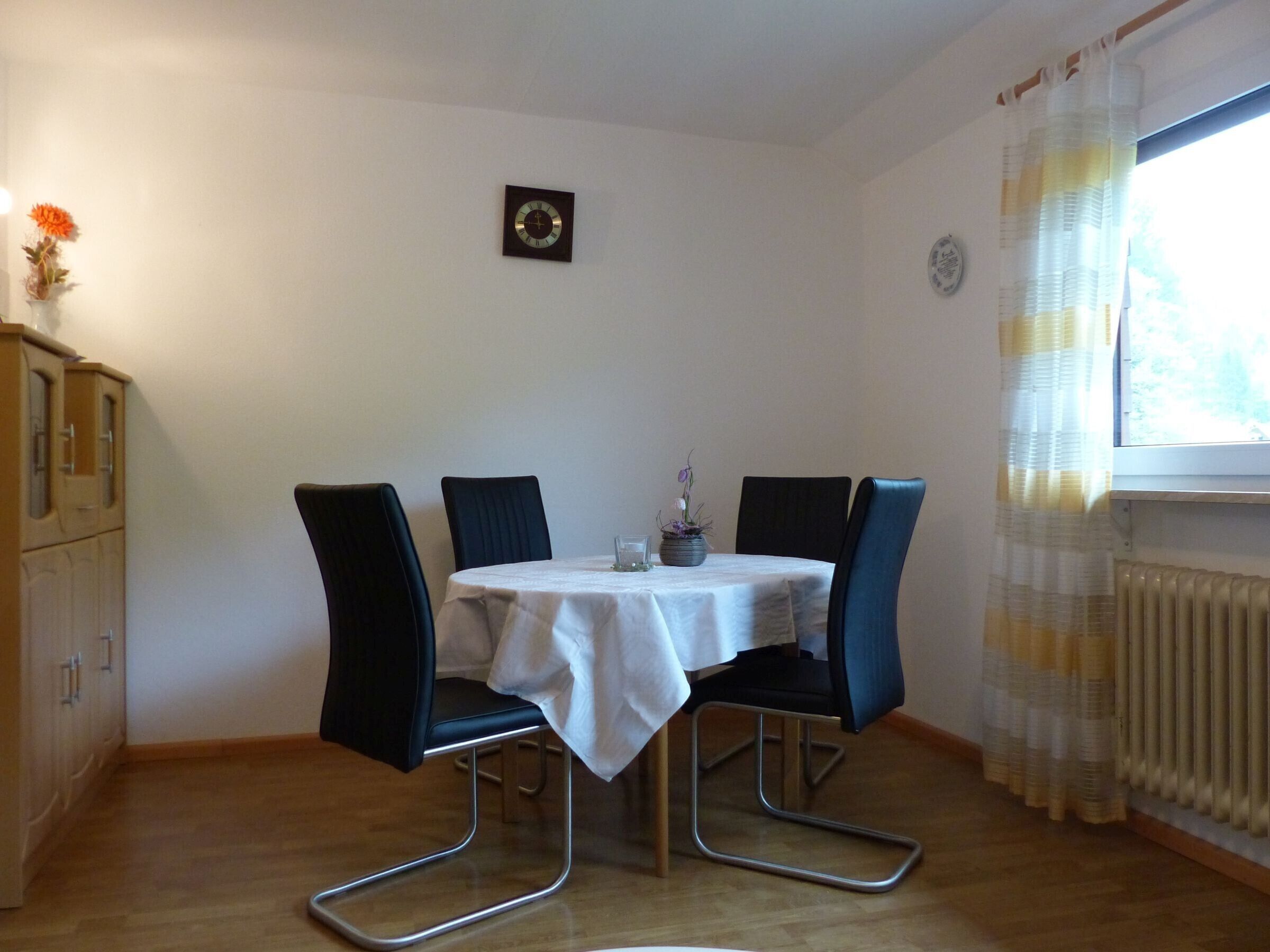 Un coin repas avec une table dressée pour quatre personnes, quatre chaises, une horloge au mur, un radiateur et une fenêtre avec des rideaux.