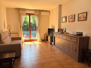 Ein Wohnzimmer mit einer Couch, einem Fernseher, einem Schrank und einem Fenster mit Vorhängen.