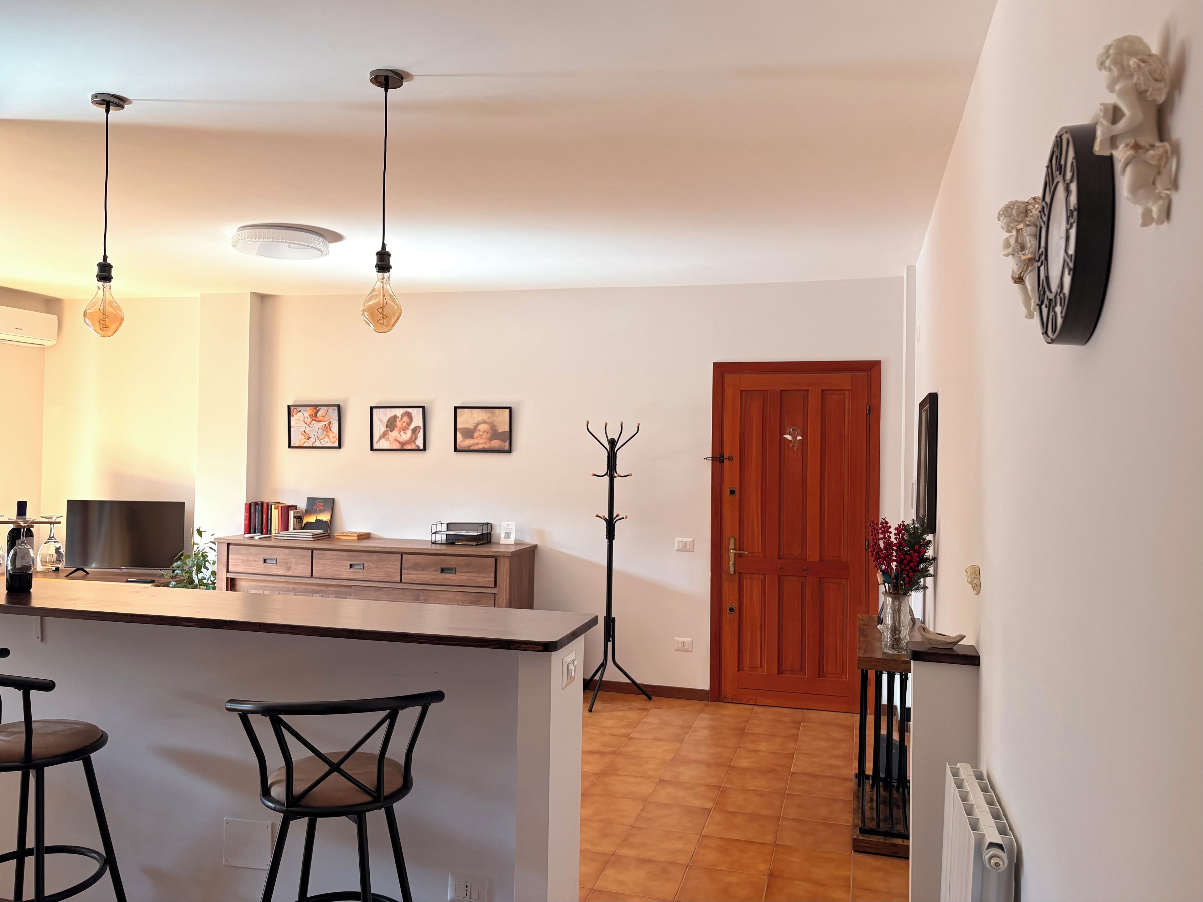 Uma cozinha com ilha central, banquetas de bar, iluminação pendente, um cabideiro e uma porta de madeira.