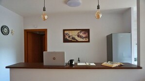 A kitchen with a laptop on the counter, a refrigerator, a painting on the wall, and a clock.