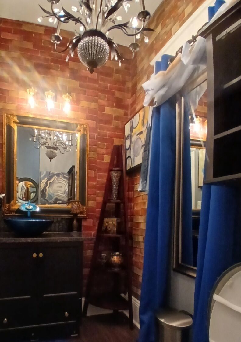 A bathroom with a brick wall, a chandelier, a mirror with a gold frame, and blue curtains.