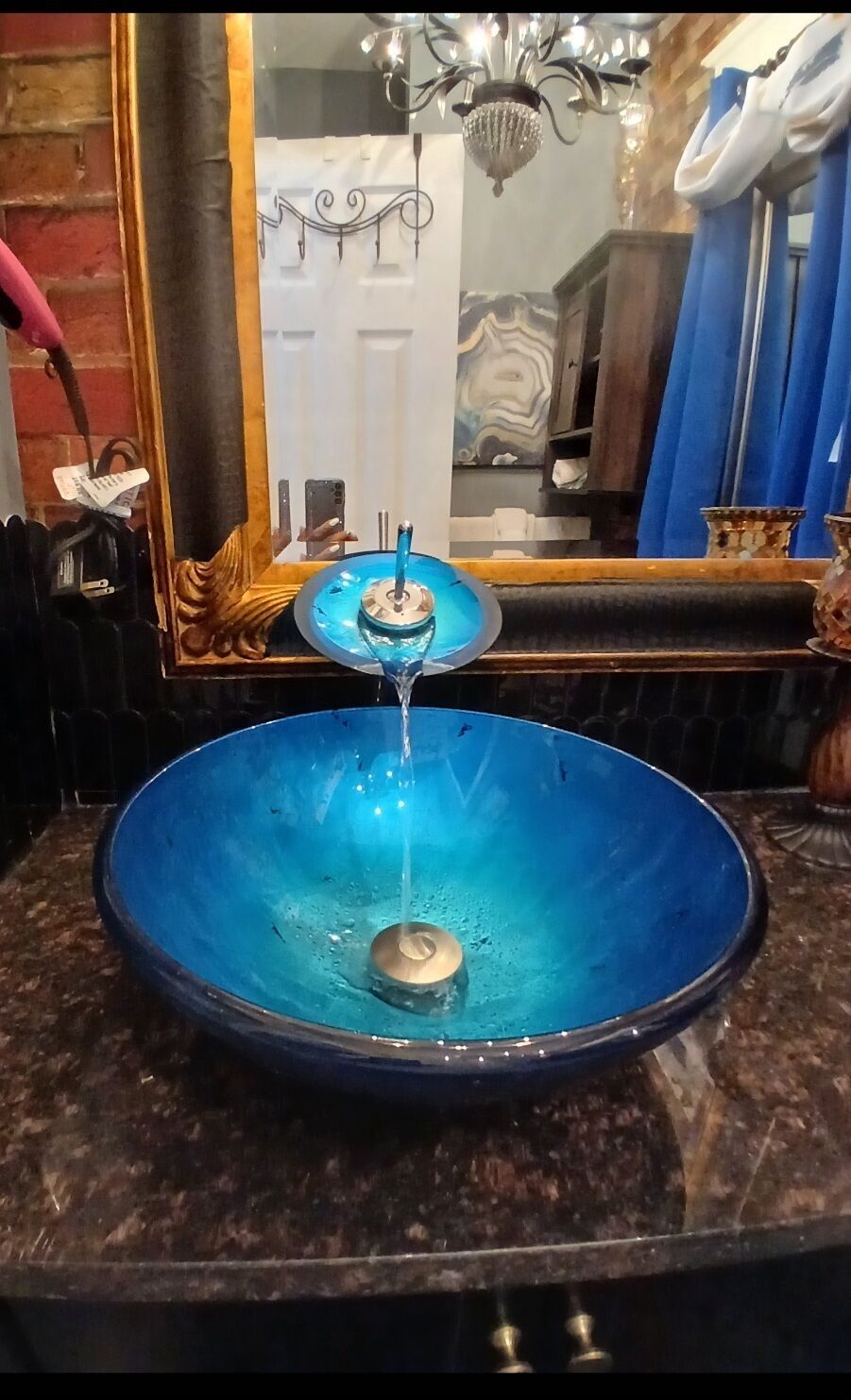 A blue glass bowl sink with a faucet, a chandelier, and a mirror reflecting a door and a blue curtain.