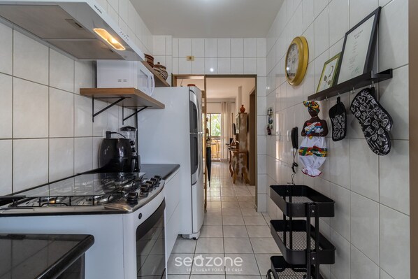 A narrow kitchen with white tiles, a gas stove, a refrigerator, and a microwave. There are black hanging racks on the wall and a small shelf above the stove. - Beautiful apt 650m From Ufsc Floripa-sc Idc0102 (Florianópolis)