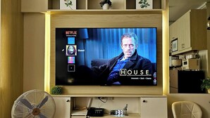 A living room with a television mounted on a wooden wall, a beige sofa, a fan, and a small plant. - Room-cozy Cebu City Condo - Heart of the (Cebu City)